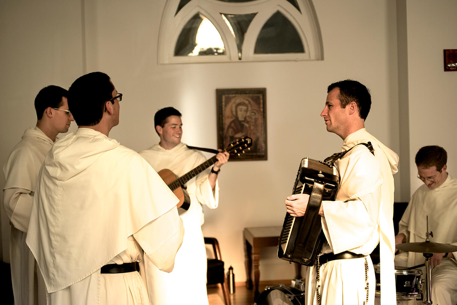 Catholic Dominican monks playing bluegrass music together in light room with arched windows.