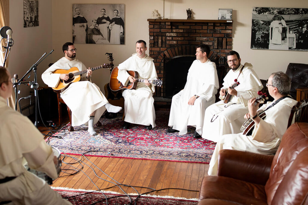 Group of Dominican brothers sitting in a living room playing bluegrass music on guitar, banjo, fiddle, mandolin.