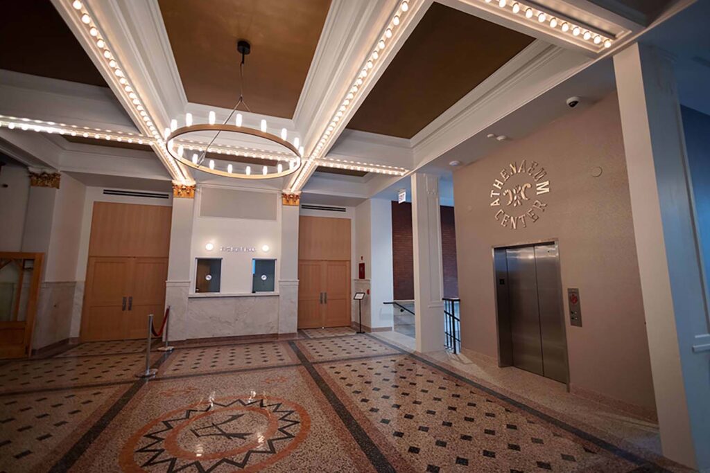 Newly renovated lobby of beautiful art deco theatre building in Lakeview Chicago with gold inset ceilings, modern round chandelier, Box office windows, and an elevator on the right.